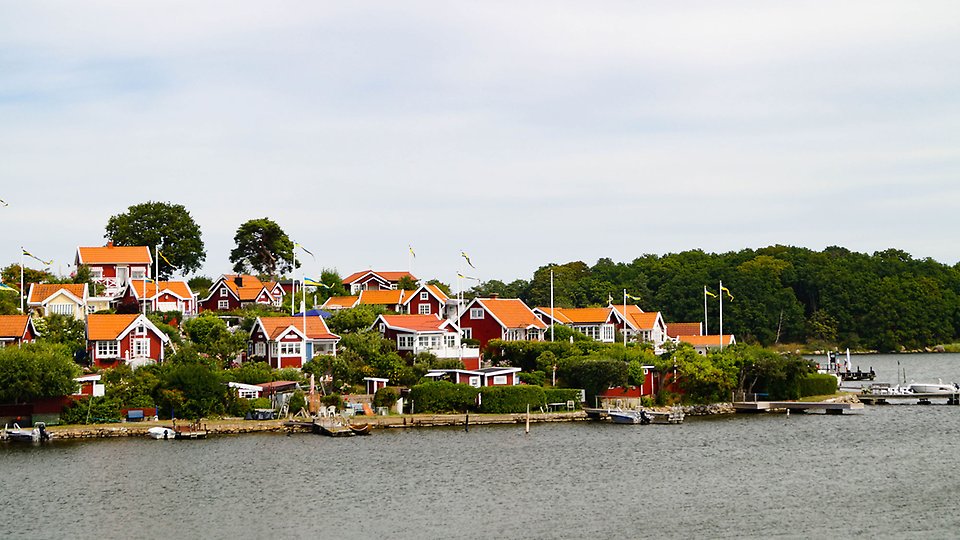 En udde med röda stugor med vita knutar. Udden grönskar i grönt, vid stranden syns bryggor och båtar. 